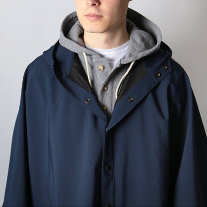 A man wearing a navy blue hooded jacket over a gray hoodie and white t-shirt, paired with an olive green cap, photographed against a plain light gray background.