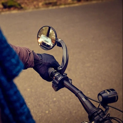 A close-up action shot of a person riding a bicycle or e-bike on an asphalt road, wearing a blue knit jacket and black gloves while gripping the handlebar with a side-view mirror attached.