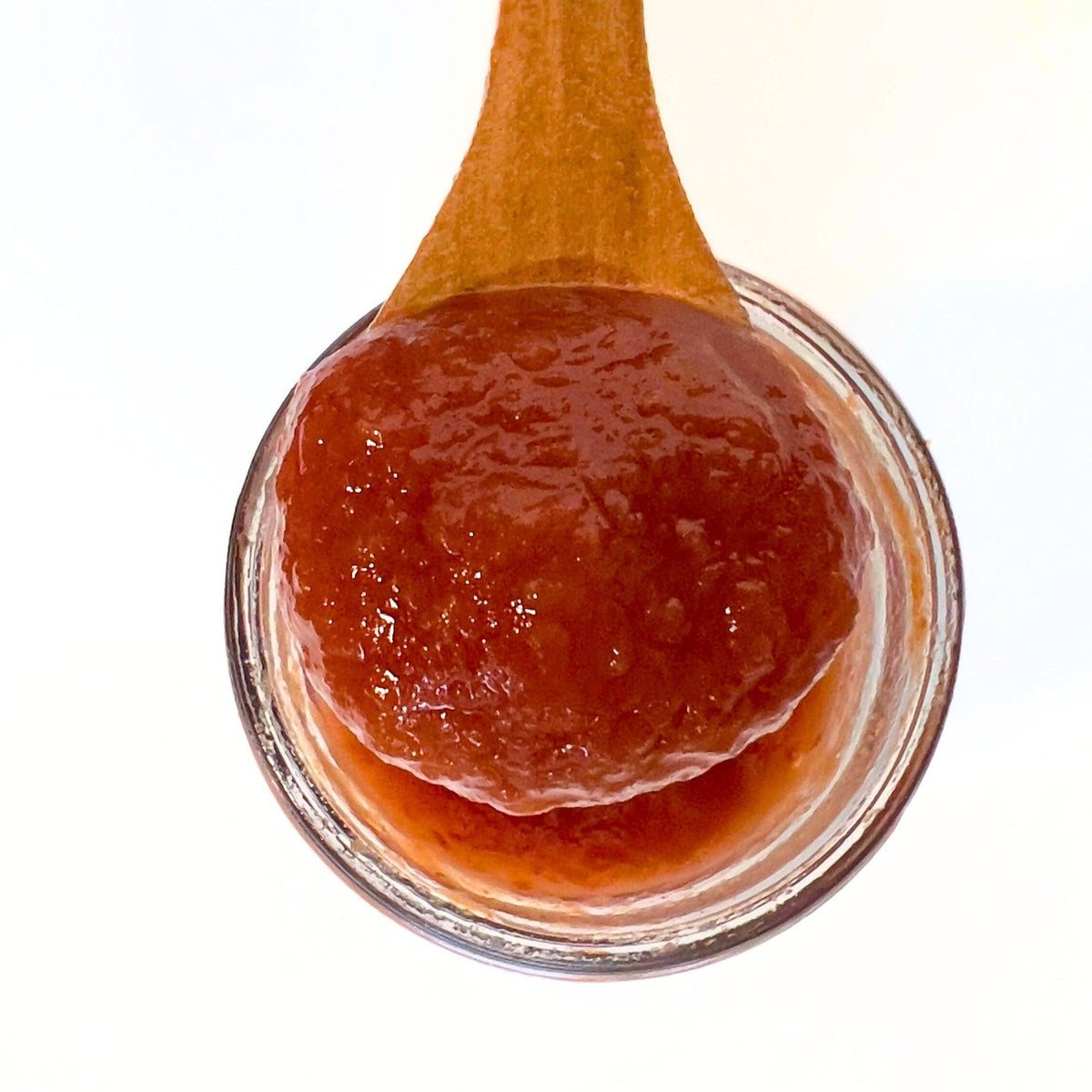 A close-up of a thick, chunky reddish-brown fruit spread or jam on a wooden spoon, held over a small clear glass bowl against a white background.