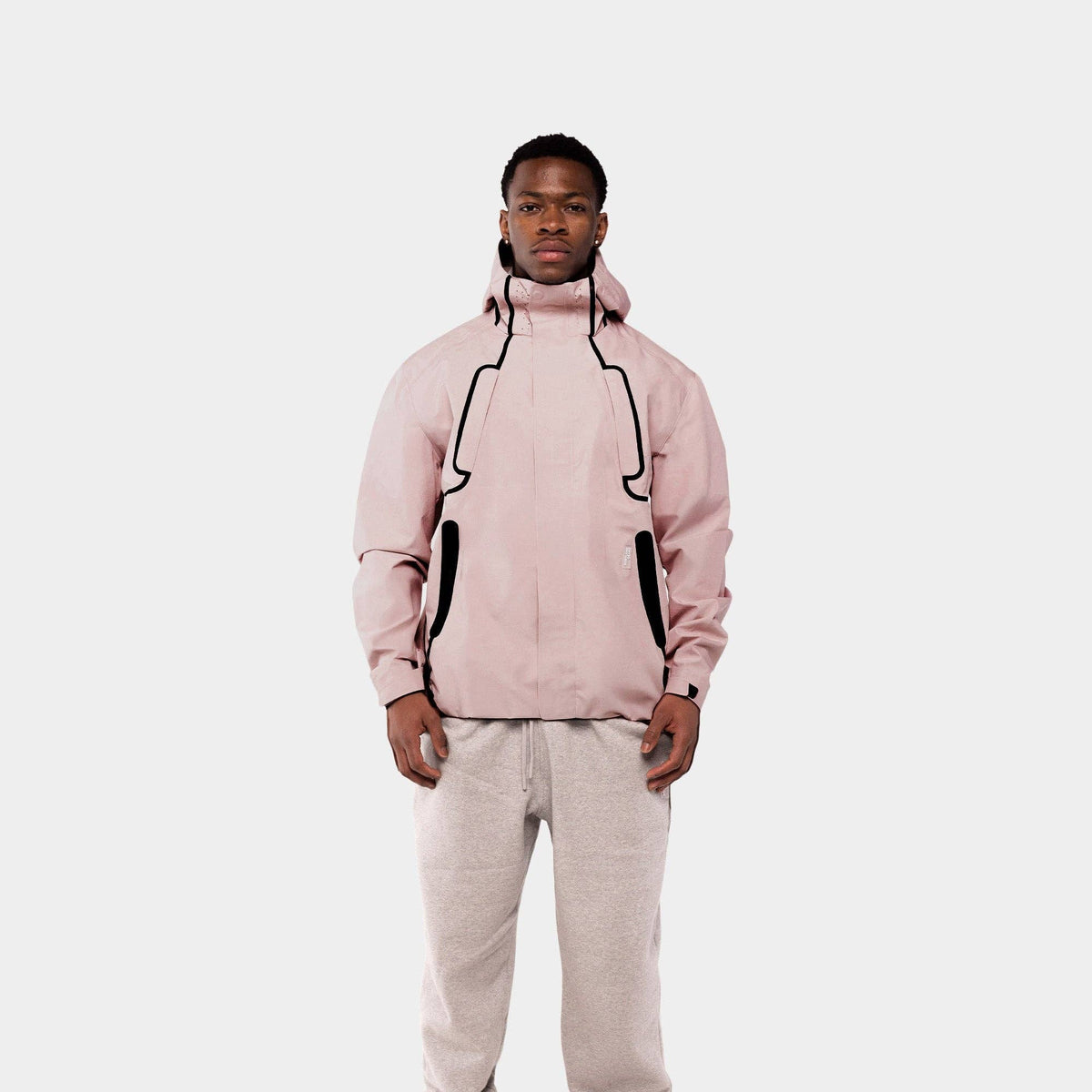 A man modeling a light pink hooded jacket with black contrast stitching and zippered pockets, paired with light gray sweatpants and white sneakers, against a solid light gray background.