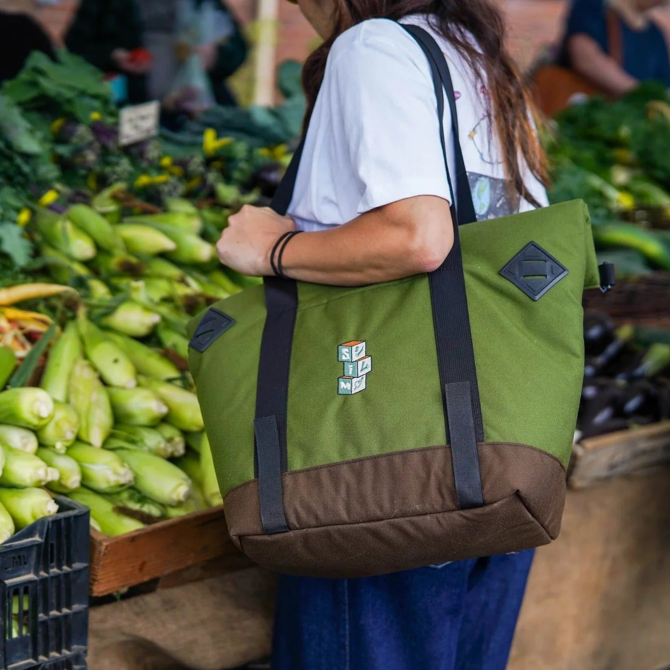 Person holding a green and brown tote bag at an outdoor market with vegetables in the background.