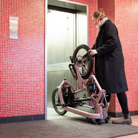 A person in a long black coat pushes a compact folding bicycle with a rear rack through a tiled elevator lobby with red mosaic walls.