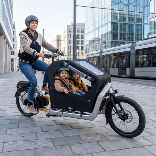 A woman rides a cargo bike in an urban setting with a child seated in the front cargo box. The scene is set on a paved plaza with modern glass buildings and a tram in the background.