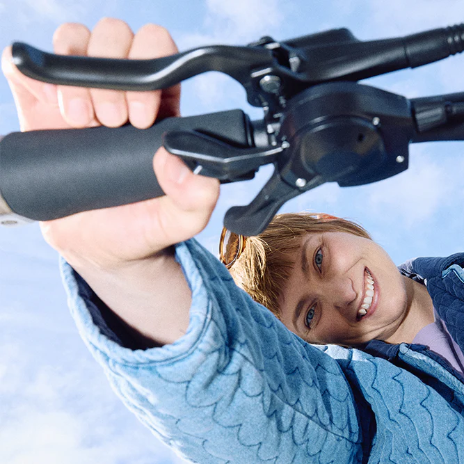 A smiling woman in a blue quilted jacket grips a black bicycle handlebar against a bright blue sky background.