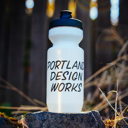 White water bottle with 'Portland Design Works' text on a rock with blurred lights in the background