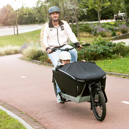 Man riding a cargo bike with a child in a black cover on a paved path in a park.