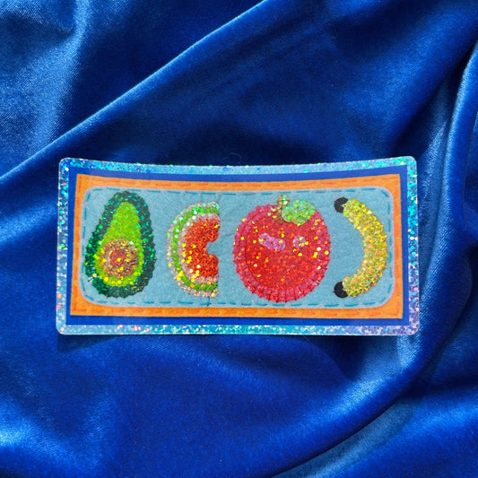 A rectangular embroidered patch featuring four colorful fruits (avocado, watermelon slice, red apple, and banana) on a glittery blue background, placed on a draped royal blue satin fabric.