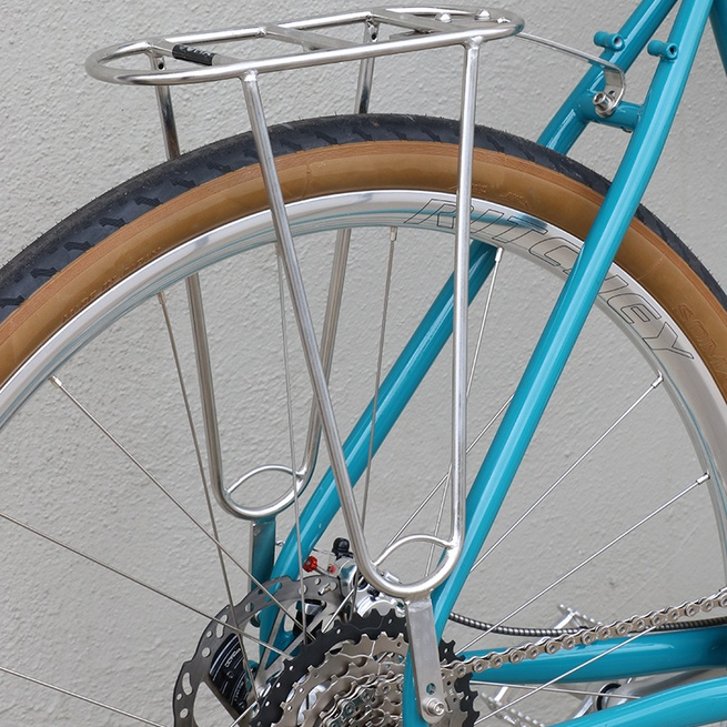 Close-up of a blue bicycle with a metal rear rack against a gray wall.
