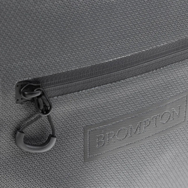 Close-up of a gray textured fabric bag with a horizontal zipper, black carabiner clip, and a debossed BROMPTON logo patch.