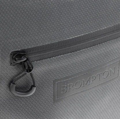 Close-up of a gray textured fabric bag with a horizontal zipper, black carabiner clip, and a debossed BROMPTON logo patch.