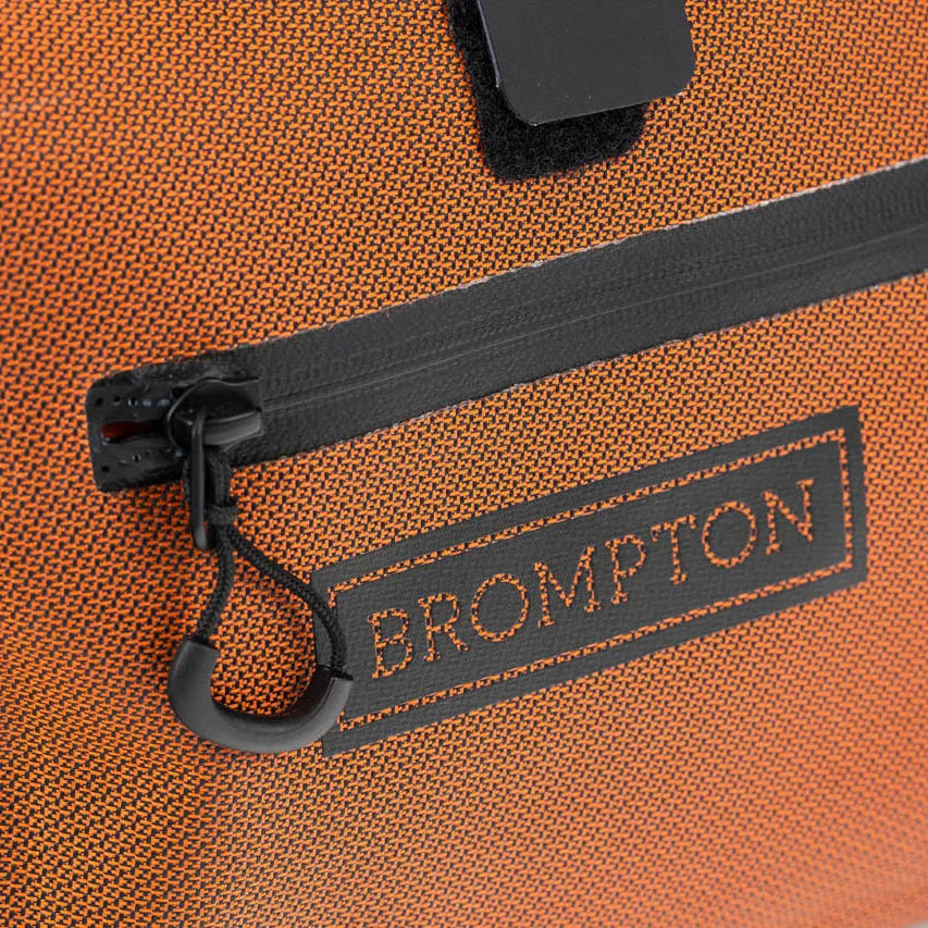 Close-up of an orange textured Brompton bag with a black zipper pocket, black carabiner clip, and a black Brompton logo patch on the front.