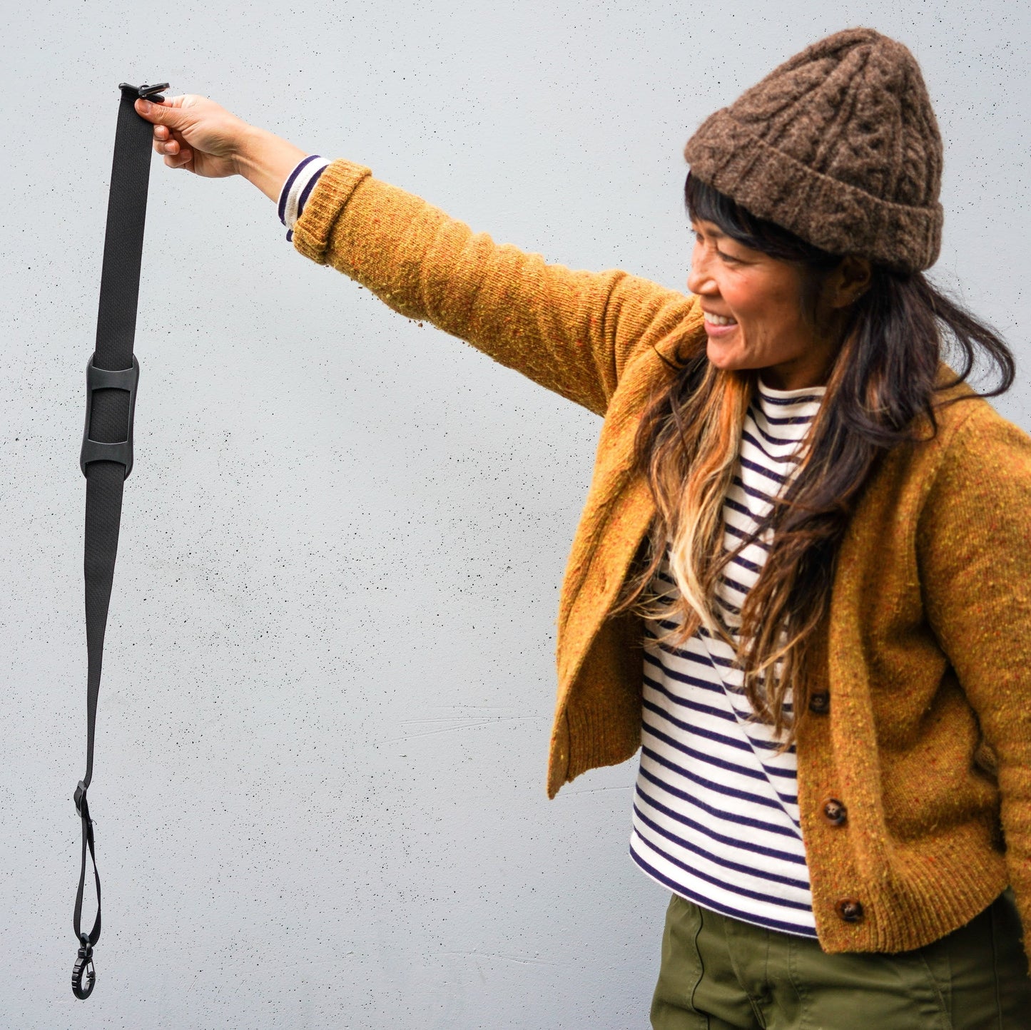 A woman wearing a brown knit beanie, mustard yellow cardigan, and striped shirt holds up a black strap with a metal clip against a light gray wall.